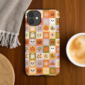 May include: A phone case with a Halloween-themed pattern. The design features a checkerboard of squares in orange, purple, green, and yellow. Each square contains a ghost, pumpkin, skull, or floral element. The case is on a wooden surface.