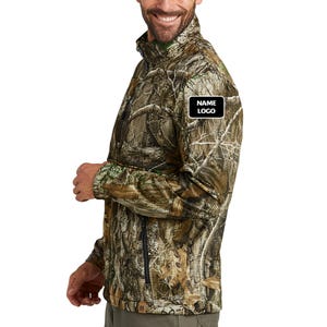 May include: A camouflage-patterned jacket with a zipper closure and a small black and white patch that reads "NAME LOGO". The jacket is a mix of brown, green, and tan colors. The person is wearing olive green pants.