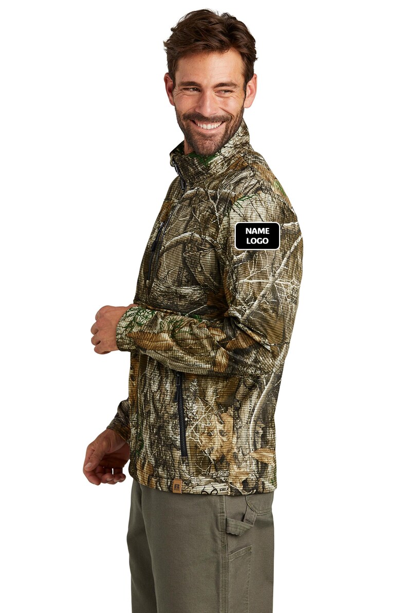 May include: A camouflage-patterned jacket with a zipper closure and a small black and white patch that reads "NAME LOGO". The jacket is a mix of brown, green, and tan colors. The person is wearing olive green pants.