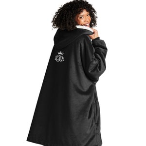 Pu&ograve; includere: Una coperta con cappuccio nera con fodera interna bianca. La coperta presenta un disegno ricamato bianco sul retro, tra cui una corona e le lettere RFS. La persona indossa leggings neri.