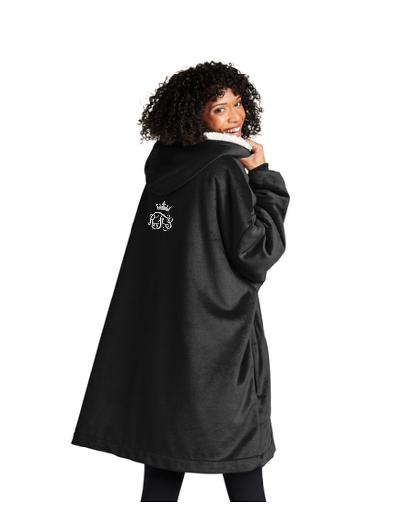 Pu&ograve; includere: Una coperta con cappuccio nera con fodera interna bianca. La coperta presenta un disegno ricamato bianco sul retro, tra cui una corona e le lettere RFS. La persona indossa leggings neri.