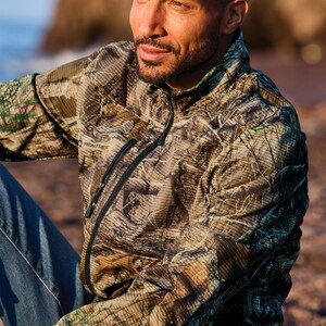 May include: A man wearing a Realtree camouflage jacket and an orange cap with a mesh back. The jacket has a black zipper and the cap has a patch that says "RUSSELL OUTDOORS". The man is wearing blue jeans.