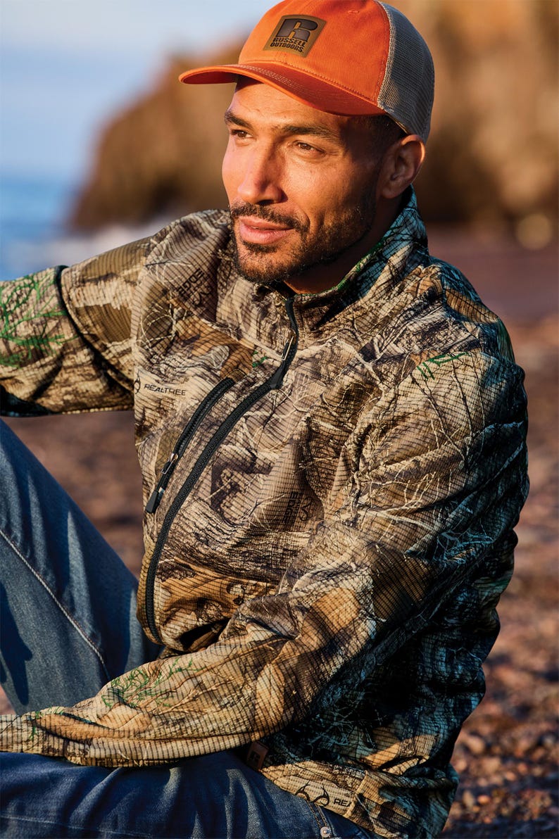 May include: A man wearing a Realtree camouflage jacket and an orange cap with a mesh back. The jacket has a black zipper and the cap has a patch that says "RUSSELL OUTDOORS". The man is wearing blue jeans.