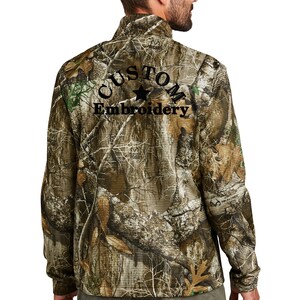 May include: A camouflage-patterned zip-up jacket with the words "CUSTOM Embroidery" embroidered in black. The jacket features a high collar and long sleeves. The overall color scheme is shades of brown, green, and gray.