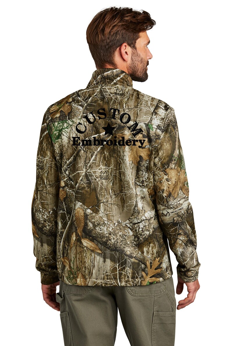 May include: A camouflage-patterned zip-up jacket with the words "CUSTOM Embroidery" embroidered in black. The jacket features a high collar and long sleeves. The overall color scheme is shades of brown, green, and gray.