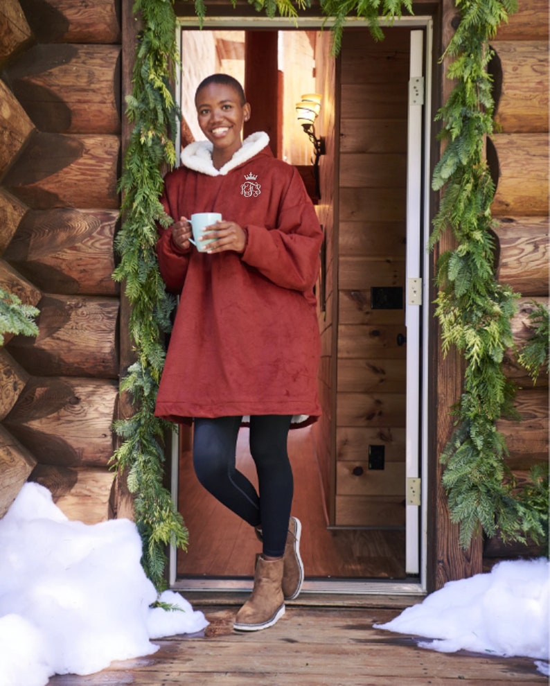 Pu&ograve; includere: Una persona indossa una coperta con cappuccio bordeaux con fodera in sherpa bianca e un'emblema a corona. La persona tiene in mano una tazza azzurra e si trova in una porta decorata con verde. Indossa leggings neri e stivali marroni.