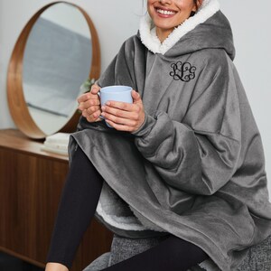 Pu&ograve; includere: Una coperta con cappuccio grigia con fodera in sherpa bianca e monogramma nero. La coperta &egrave; indossata sopra leggings neri. La persona tiene in mano una tazza azzurra.