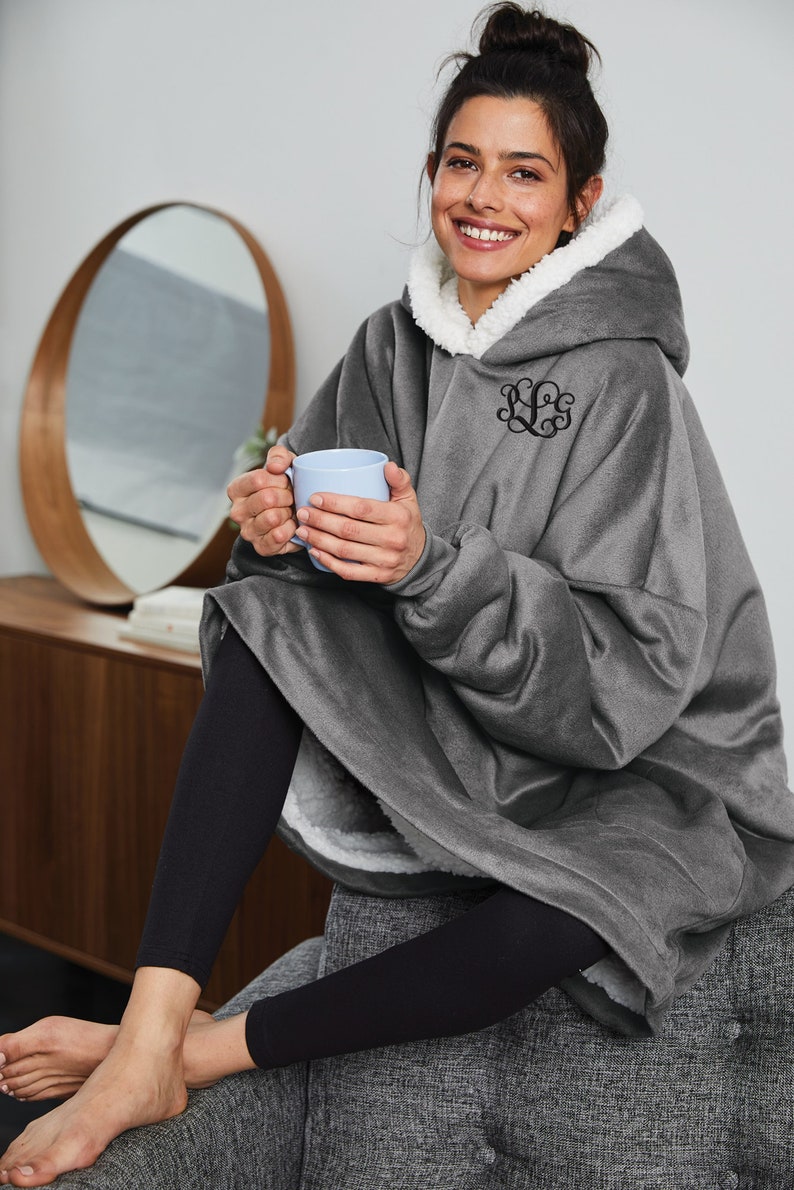 Pu&ograve; includere: Una coperta con cappuccio grigia con fodera in sherpa bianca e monogramma nero. La coperta &egrave; indossata sopra leggings neri. La persona tiene in mano una tazza azzurra.
