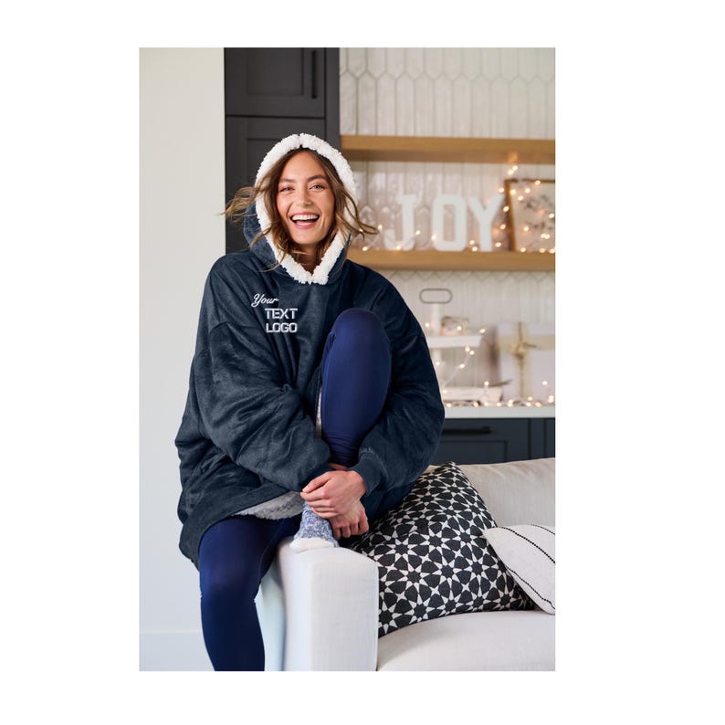 Pu&ograve; includere: Una coperta con cappuccio oversize blu navy con cappuccio foderato in sherpa bianco. La coperta ha il testo "Your TEXT LOGO" ricamato sul davanti. La persona indossa leggings blu navy.