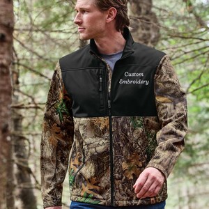 May include: A full-zip jacket with a black upper section and camouflage print sleeves and lower body. The jacket has the words "Custom Embroidery" embroidered on the chest. The person is wearing blue jeans.