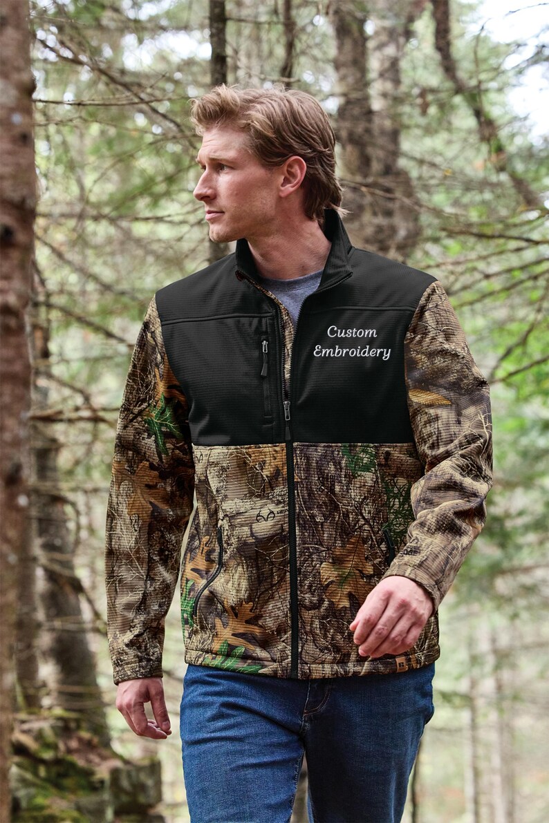 May include: A full-zip jacket with a black upper section and camouflage print sleeves and lower body. The jacket has the words "Custom Embroidery" embroidered on the chest. The person is wearing blue jeans.