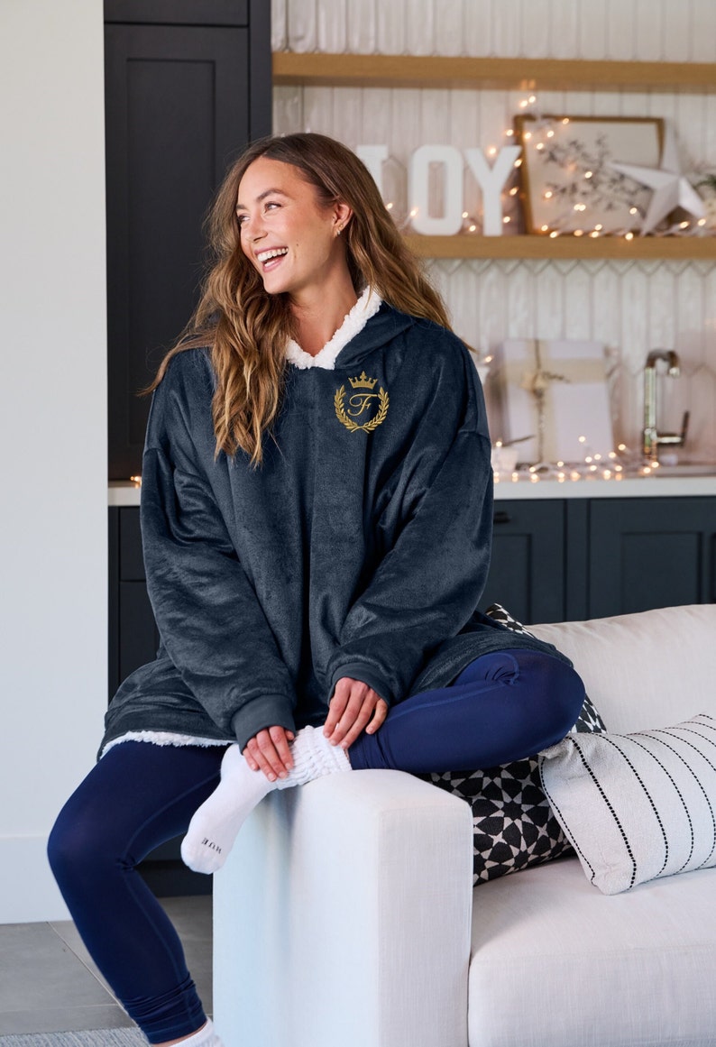 Pu&ograve; includere: Una coperta con cappuccio oversize blu navy con fodera in sherpa bianca e un emblema dorato ricamato. La persona indossa leggings blu navy e calzini bianchi. La coperta &egrave; morbida e confortevole.
