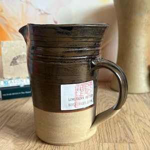May include: A ceramic pitcher with a dark brown glazed upper section and a natural tan unglazed base. The pitcher features a side handle, a pouring spout, and a white rectangular label reading Jeremy Leach Lowerdown Pottery Bovey Tracey Devon.