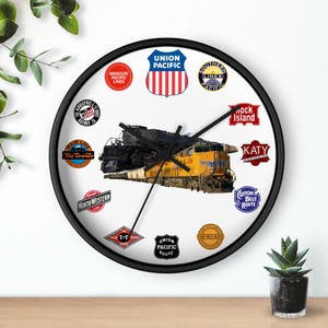 May include: A round wall clock with a black frame. The clock face features a yellow and black train image, surrounded by various railroad logos. The clock hands are black.