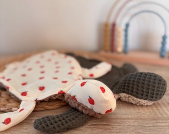 May include: A soft turtle-shaped security blanket featuring a cream-colored fabric with a red apple print on the head and top shell, contrasted with a dark olive green waffle-textured fabric on the underside and flippers, with a plush texture.
