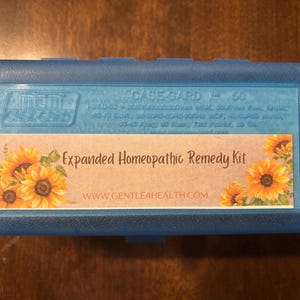 Puede incluir: Un estuche de almacenamiento rectangular de plástico azul con una etiqueta que presenta ilustraciones de girasoles amarillos y el texto Expanded Homeopathic Remedy Kit y WWW.GENTLE4HEALTH.COM. La tapa tiene grabado MTM CASE-GARD 50.