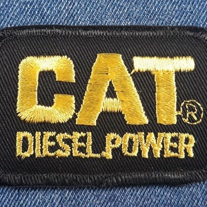 May include: Black embroidered patch with the word "CAT" in gold lettering and the words "DIESEL POWER" in gold lettering below. The "CAT" logo has a registered trademark symbol.