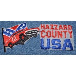 May include: A red, white, and blue embroidered patch with a car and a Confederate flag. The text reads "Hazzard County USA".
