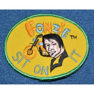 May include: A yellow and green embroidered patch with a cartoon image of a man riding a motorcycle. The text "Fonzie's TM Sit On It" is embroidered around the image.