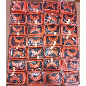 May include: A box filled with numerous rectangular patches. Each patch features the words "TRANS AM" and a stylized orange bird emblem on a black background, surrounded by an orange border. The patches are individually packaged in clear plastic.