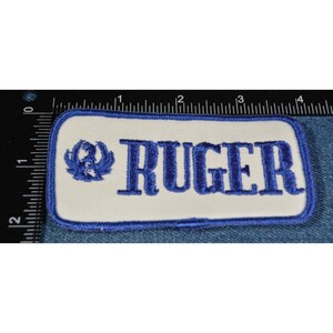 May include: A white rectangular patch with blue embroidered text that reads "RUGER". A blue embroidered logo of a stylized eagle is to the left of the text.