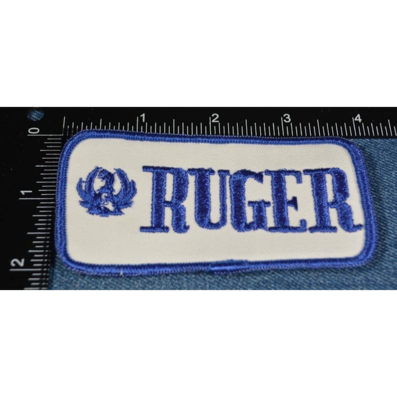 May include: A white rectangular patch with blue embroidered text that reads "RUGER". A blue embroidered logo of a stylized eagle is to the left of the text.