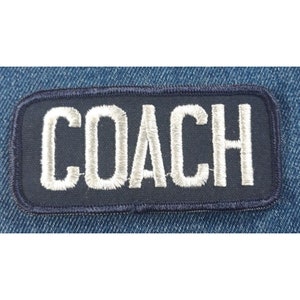 May include: A rectangular navy blue embroidered patch with the word "COACH" in white, stitched lettering. The patch has a dark blue border and is set against a denim background.