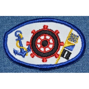 May include: A white oval patch with a blue border, featuring a red and black ship's wheel, a blue anchor with a gold rope, and a yellow and blue striped flag.