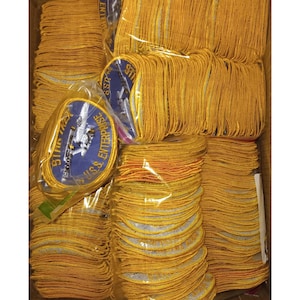 Puede incluir: Una caja llena de montones de parches bordados amarillos y dorados. Algunos parches son redondos con un centro azul y el texto "U.S.S. ENTERPRISE" y "STAR TREK". Los parches están en varias etapas de embalaje.