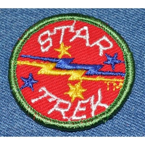 May include: A red, white, and blue embroidered patch with the words "STAR TREK" and a lightning bolt design. The patch is surrounded by a green border.