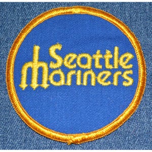 May include: A circular patch featuring the words "Seattle Mariners" in yellow script on a blue background. The patch is bordered by a gold-colored stitched edge. The patch is likely for a baseball team.