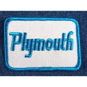 May include: A rectangular patch with the word "Plymouth" embroidered in teal blue on a white background. The patch has a teal blue border and is set against a textured blue fabric.