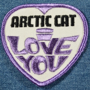 May include: White heart-shaped patch with purple embroidered text that reads "LOVE YOU" and a purple stylized microphone. The text "ARCTIC CAT" is embroidered in black above the microphone.