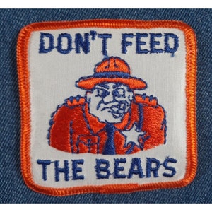 May include: A white patch with an orange border, featuring a cartoon bear wearing a ranger hat and uniform. The text "DON'T FEED THE BEARS" is written in blue lettering.