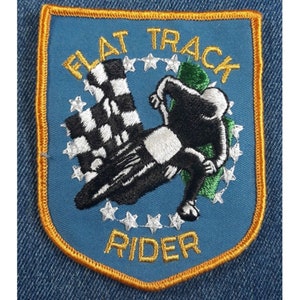 May include: A blue and gold embroidered patch with a checkered flag and a rider on a motorcycle. The text "FLAT TRACK RIDER" is embroidered in gold.