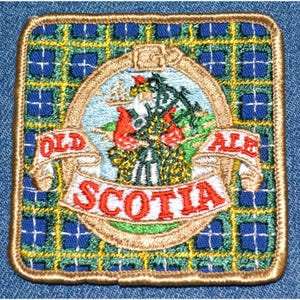 May include: Square embroidered patch with a gold border and a tartan background in blue, green, and yellow. The patch features a central design with the words "OLD" and "ALE" on banners, and "SCOTIA" in red. The design includes a floral and landscape scene.