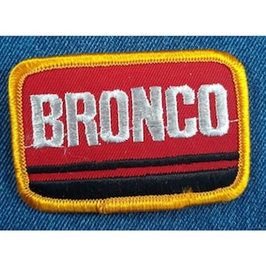 May include: A red embroidered patch with a yellow border and black stripes. The patch says "BRONCO" in white lettering.