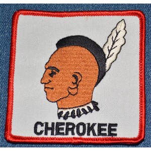 May include: Embroidered patch featuring a profile of a Native American with a black mohawk and a feather. The patch has a white background with a red border and the word "CHEROKEE" in black.