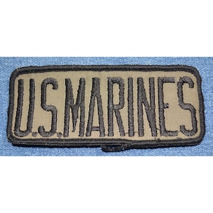 May include: A tan fabric patch with black embroidered text that reads "U.S. MARINES".