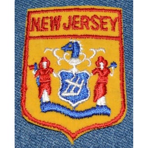 May include: A yellow felt patch with a red border, depicting the state seal of New Jersey. The seal features a horse's head, two figures holding objects, and a shield with a blue background and white accents. The text "NEW JERSEY" is embroidered in red at the top of the patch.