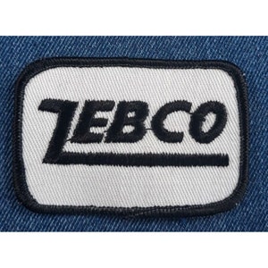 May include: White rectangular patch with black embroidered text that reads "ZEBCO" with a black line underneath.