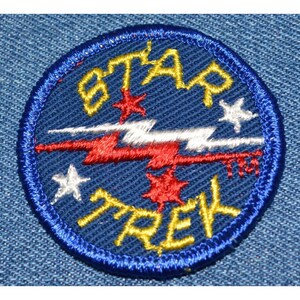 May include: A blue and yellow embroidered patch with the words "STAR TREK" in a stylized font. The patch features a red, white, and blue lightning bolt design with four stars.
