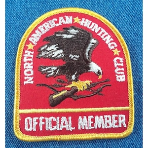 May include: Red embroidered patch with a yellow border. The patch features a brown eagle with white markings, holding a rifle in its talons. The text "NORTH AMERICAN HUNTING CLUB" is written in a yellow arc around the eagle. The text "OFFICIAL MEMBER" is written in white below the eagle.