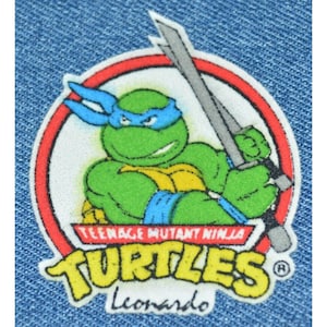 May include: A denim patch featuring Leonardo from Teenage Mutant Ninja Turtles. The green turtle is wearing a blue mask and holding a silver sword. The patch has a red and white border with the words "Teenage Mutant Ninja Turtles" and "Leonardo".