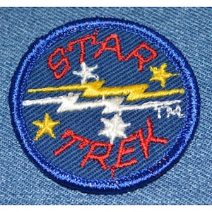 May include: A blue circular embroidered patch with the text "STAR TREK" in red, "STAR" in red, and "TM" in white. The patch features a white lightning bolt with yellow accents and five stars, two yellow and three white.