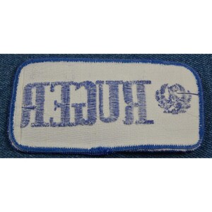 May include: A white fabric patch with blue embroidered text that reads "RUGER" and a blue embroidered logo of a stylized eagle.