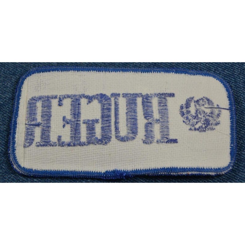 May include: A white fabric patch with blue embroidered text that reads "RUGER" and a blue embroidered logo of a stylized eagle.