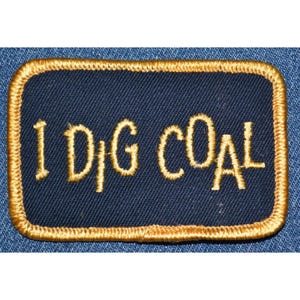 May include: Embroidered patch with the phrase "I DIG COAL" in gold thread on a navy blue background. The patch has a gold border and is rectangular in shape. The patch is likely for a work uniform or a collector's item.