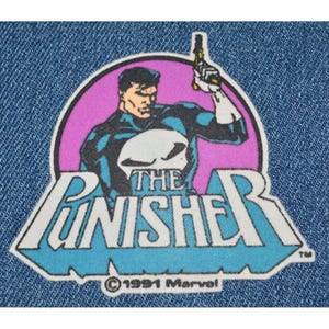 May include: A blue and white embroidered patch featuring The Punisher, a comic book character. The patch has a pink circle with a black and white illustration of The Punisher holding a gun. The text "The Punisher" is in white letters on a blue background. The text "© 1991 Marvel" is in white letters on a blue background.