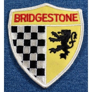 May include: A white, yellow, and black embroidered patch with the word "BRIDGESTONE" in red lettering. The patch features a checkered pattern and a black lion.